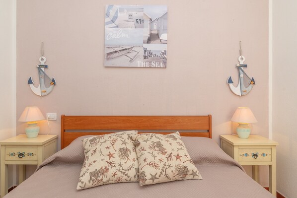 Un dormitorio con cabecera de madera, dos mesitas de noche con lámparas, cama con manta color beige y almohadas estampadas con estrellas, y un cuadro colgado en la pared que representa una playa.