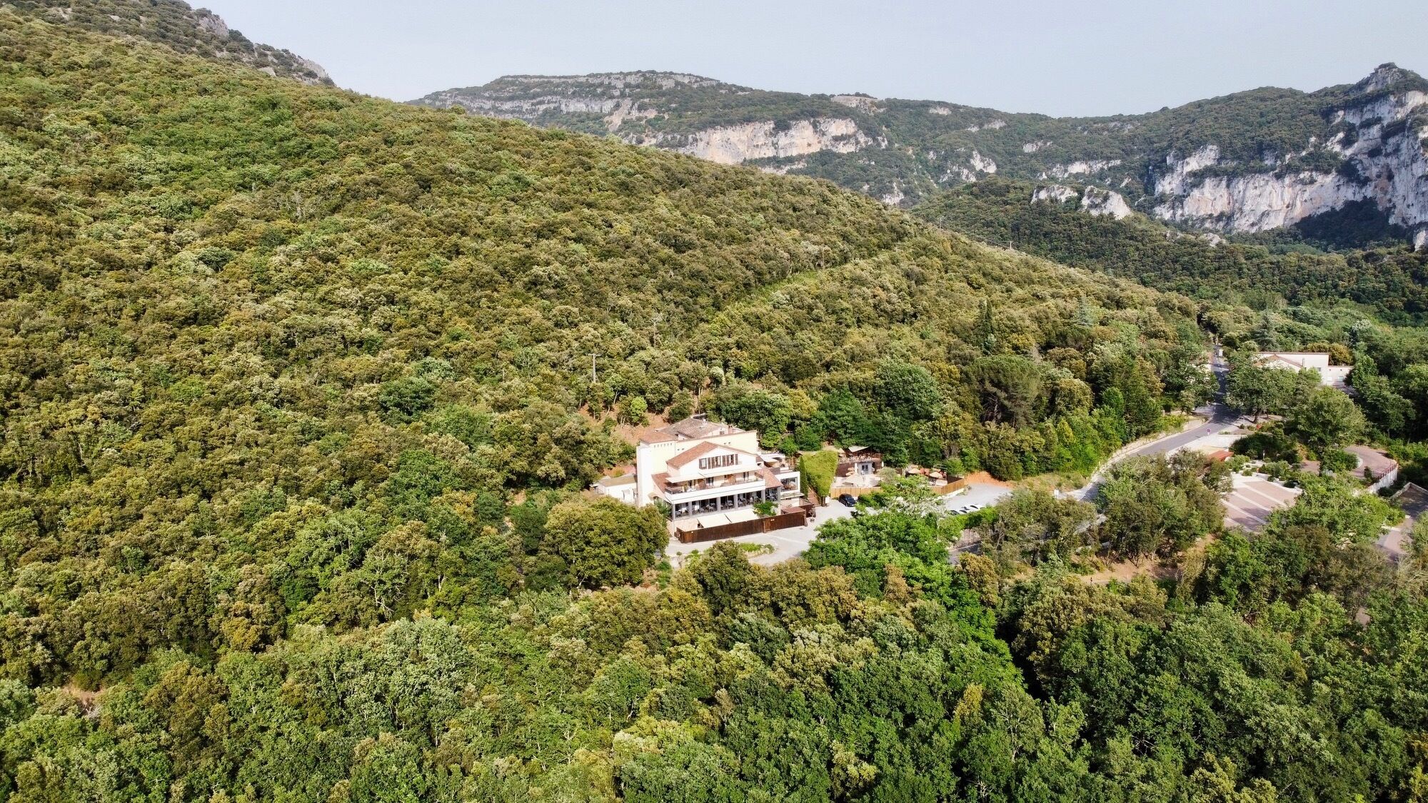 A two-story building with a flat roof, surrounded by dense forest, set against a backdrop of hills.