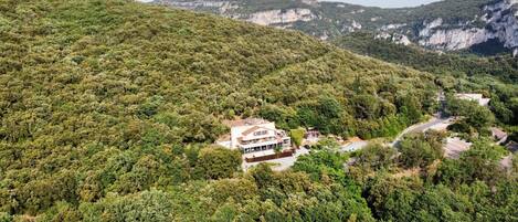 A two-story building with a flat roof, surrounded by dense forest, set against a backdrop of hills.