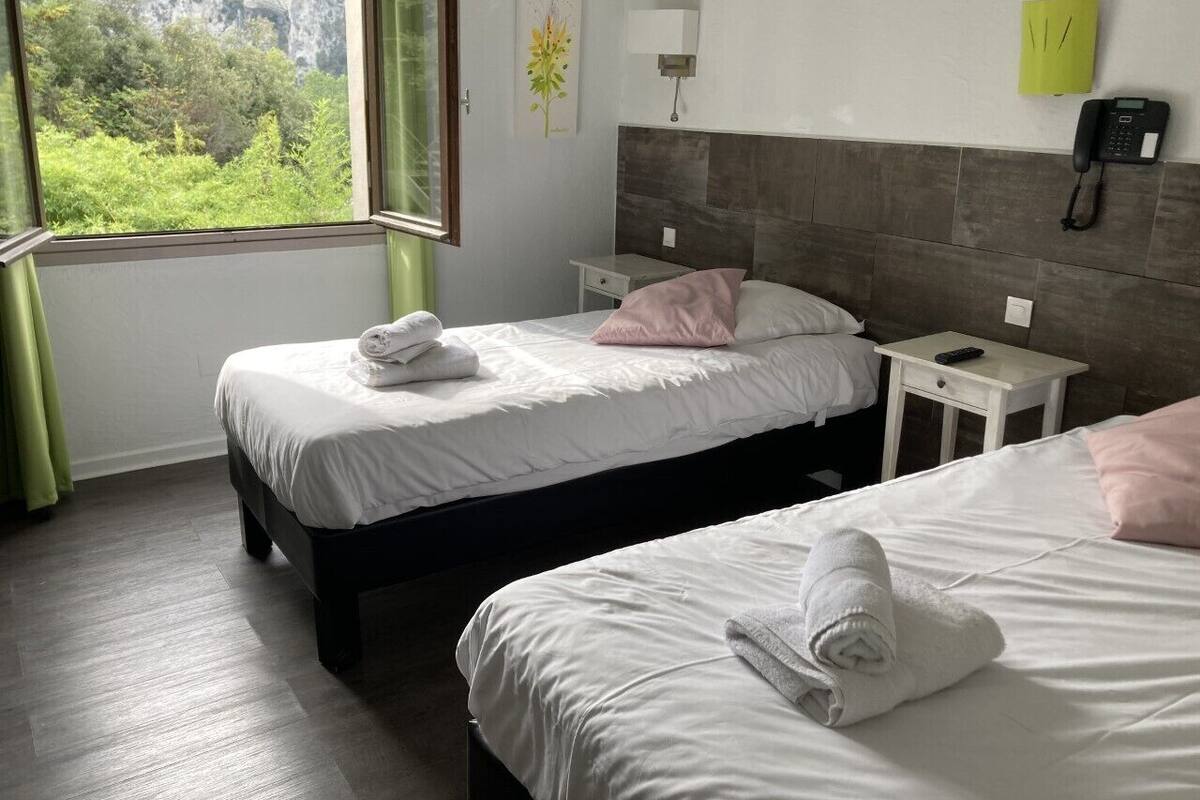 Une chambre d’hôtel avec deux lits, une table de chevet avec une lampe, un téléphone et une fenêtre donnant sur un paysage verdoyant et montagneux.