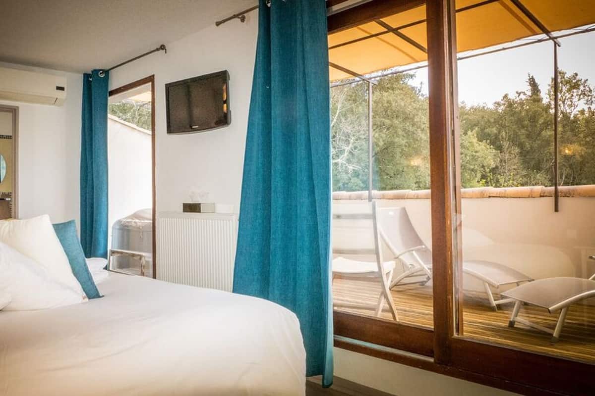 Une chambre avec un grand lit, une fenêtre avec des rideaux bleus, une petite table avec une boîte de mouchoirs et une chaise placée sur un balcon.