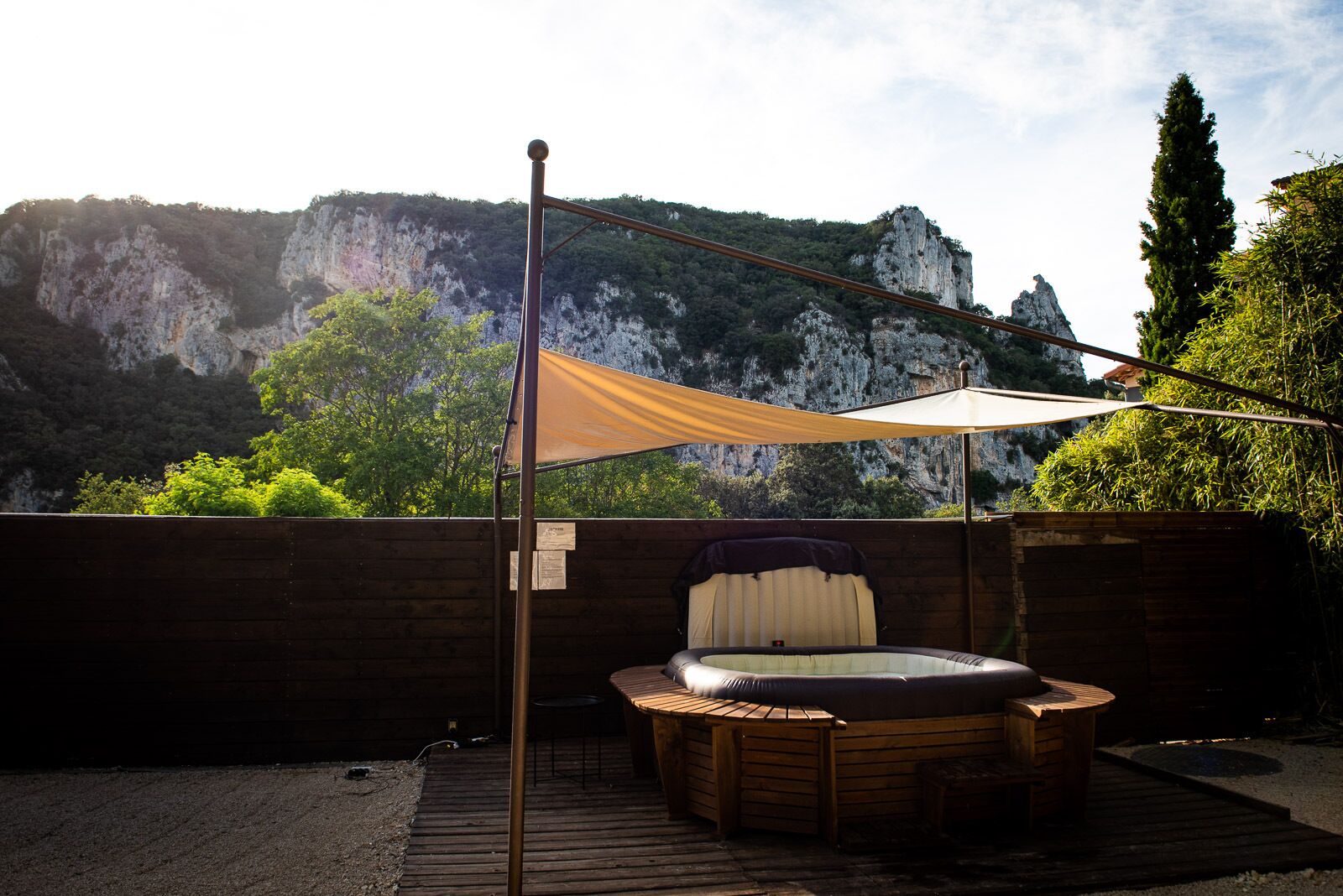 A hot tub with a wooden deck and a canopy, set against a backdrop of rocky cliffs and greenery.