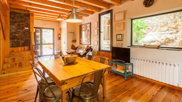Una mesa de comedor de madera con sillas, un televisor sobre un mueble bajo y una ventana con vistas a un muro de piedra.