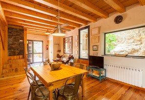 Ein Holztisch mit Stühlen, ein Fernseher auf einem Schrank und ein Fenster mit Blick auf eine Steinmauer.