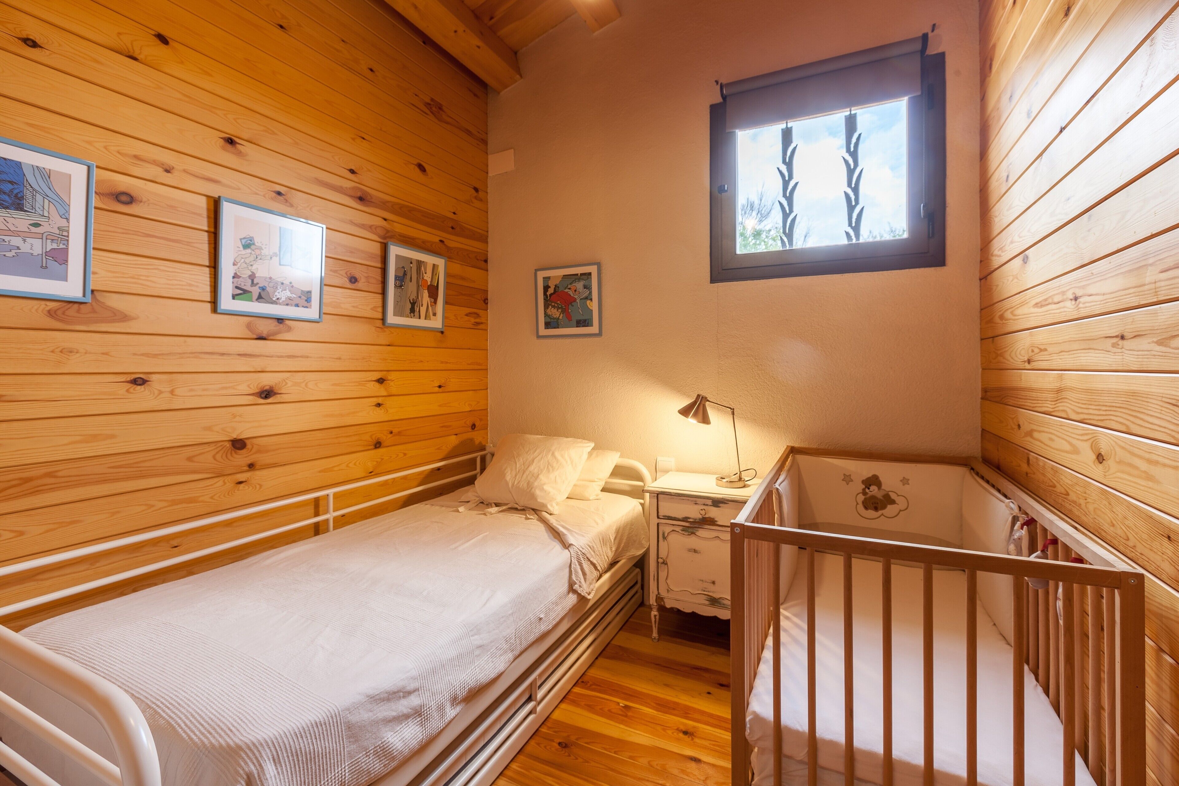 A wooden-paneled room with a single bed, a nightstand, a crib, and framed pictures on the wall.