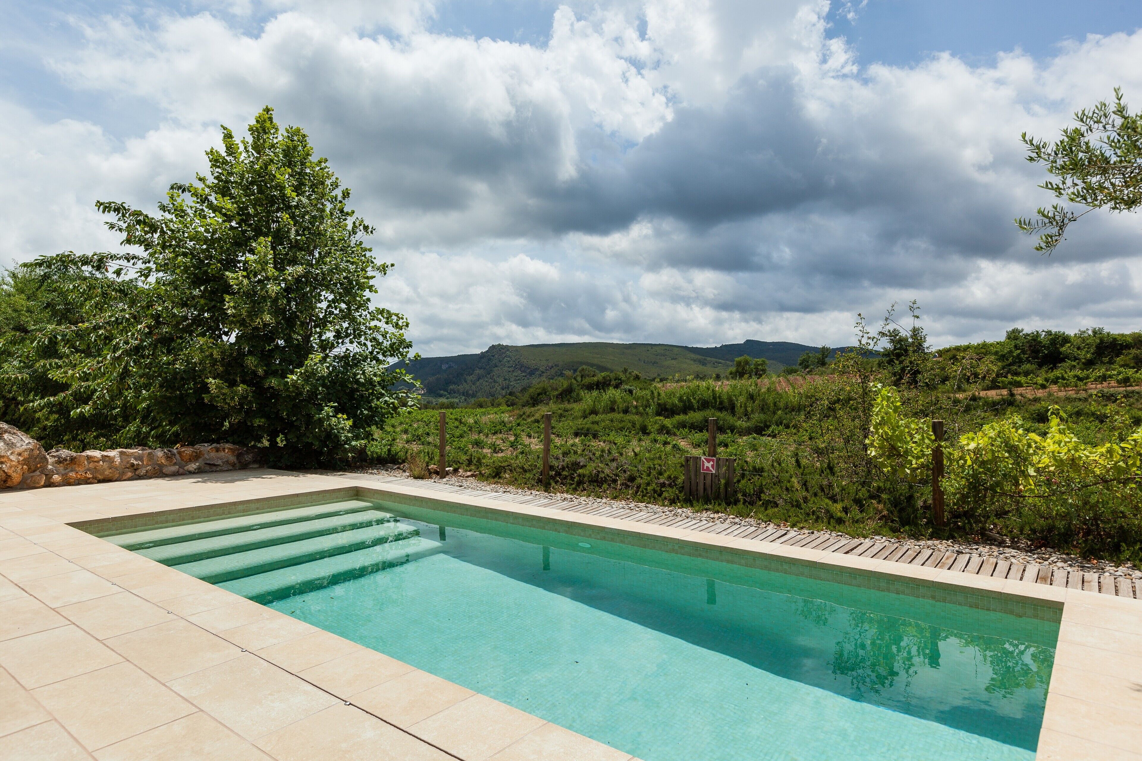 Una piscina rectangular de agua cristalina, rodeada por un borde de piedra, con un exuberante follaje y colinas al fondo bajo un cielo nublado.