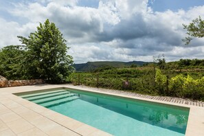 Ein rechteckiger Pool mit klarem Wasser, umgeben von einer Steinmauer, vor einer Kulisse aus üppiger Vegetation und Hügeln unter bewölktem Himmel.