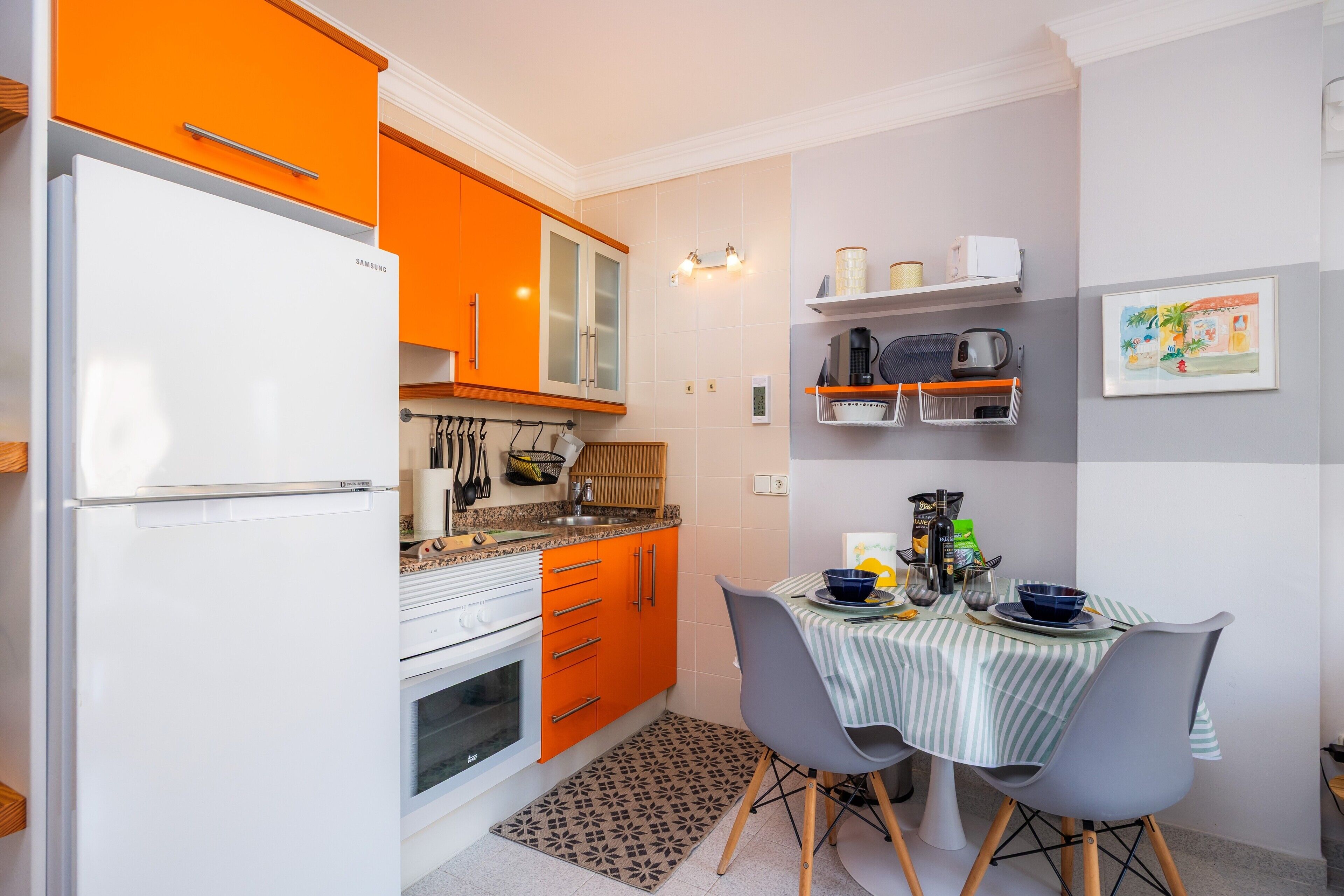 Une cuisine compacte équipée d’un réfrigérateur, d’un four et de meubles de couleur orange. On y trouve une table à manger avec des chaises et un tapis à motifs.