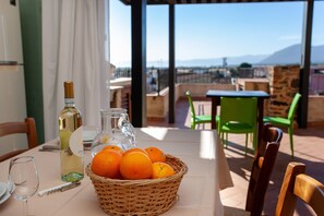 A table with a basket of oranges, a wine bottle, and a glass on a terrace with chairs and a view of the city.