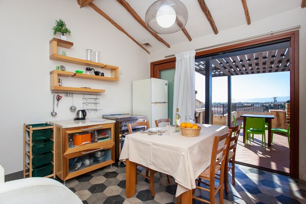 A kitchen with wooden shelves, a refrigerator, a dining table with chairs, and a view through a glass door.