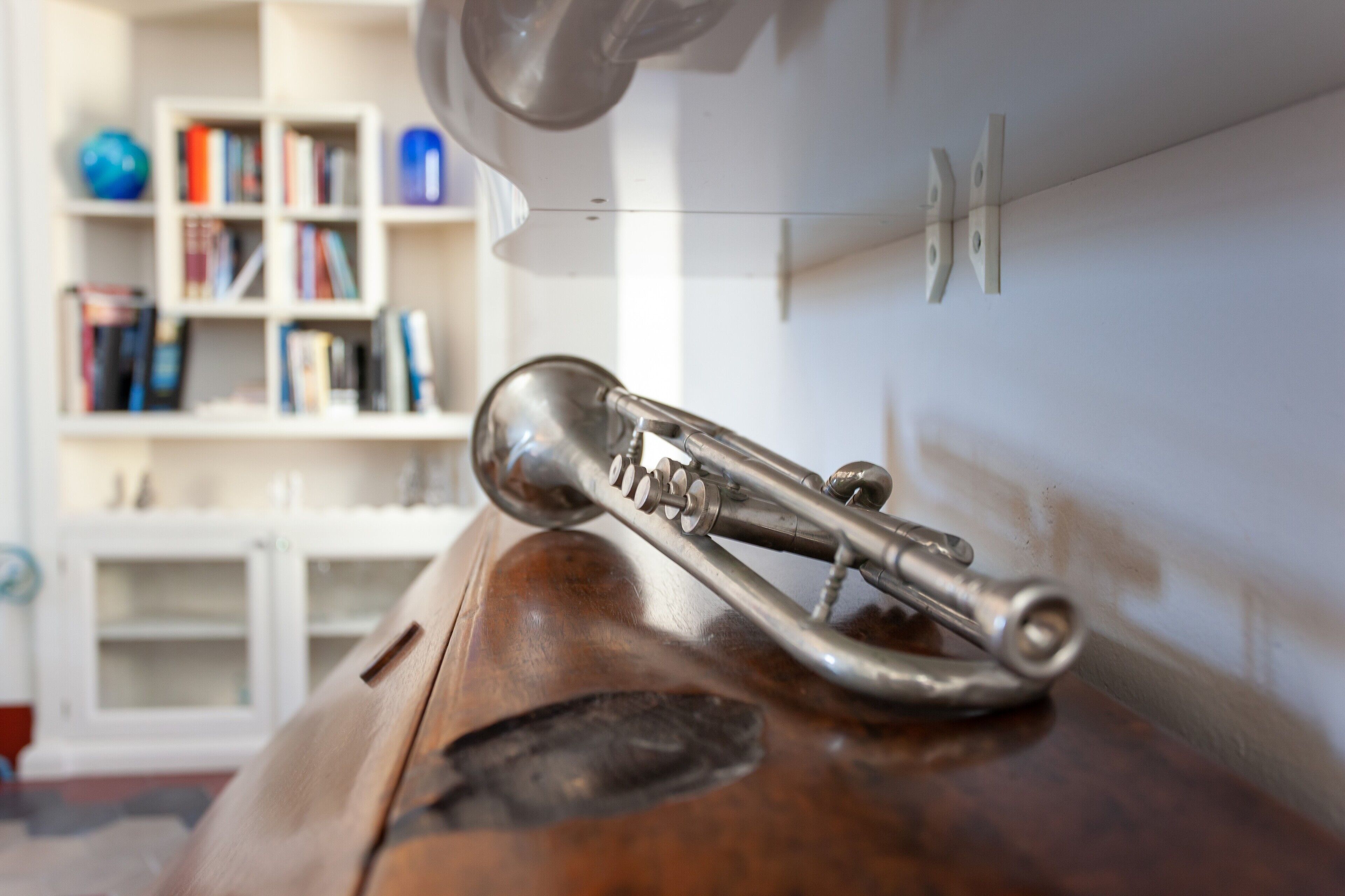 A brass trumpet on a wooden surface, a white shelf with books, and a white cabinet in the background.