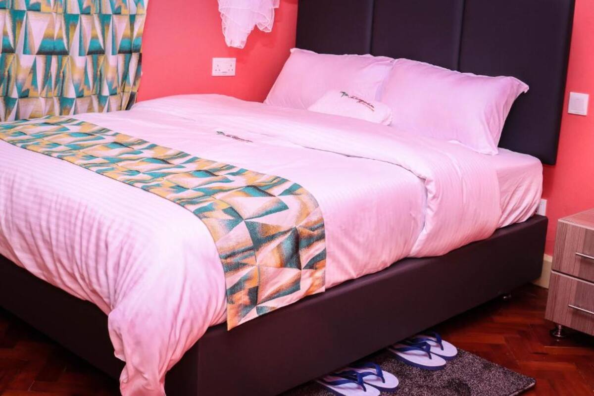 Una cama con un edredón estampado con motivos geométricos, un cabecero oscuro y un par de chanclas sobre una alfombra.