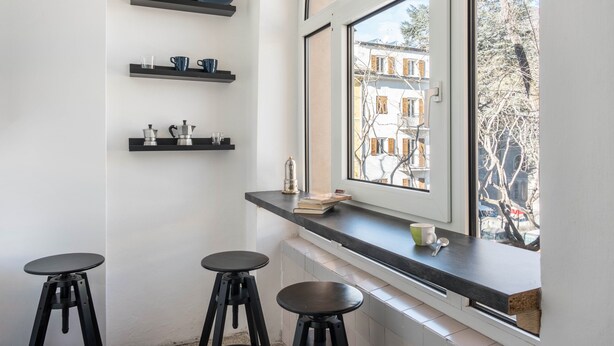 Una cocina con taburetes negros, una ventana con vistas a edificios y estantes con utensilios de cocina.