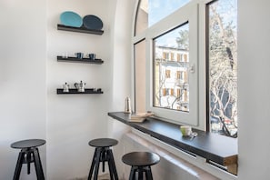 A kitchen with black stools, a window with a view of buildings, and shelves with kitchenware.