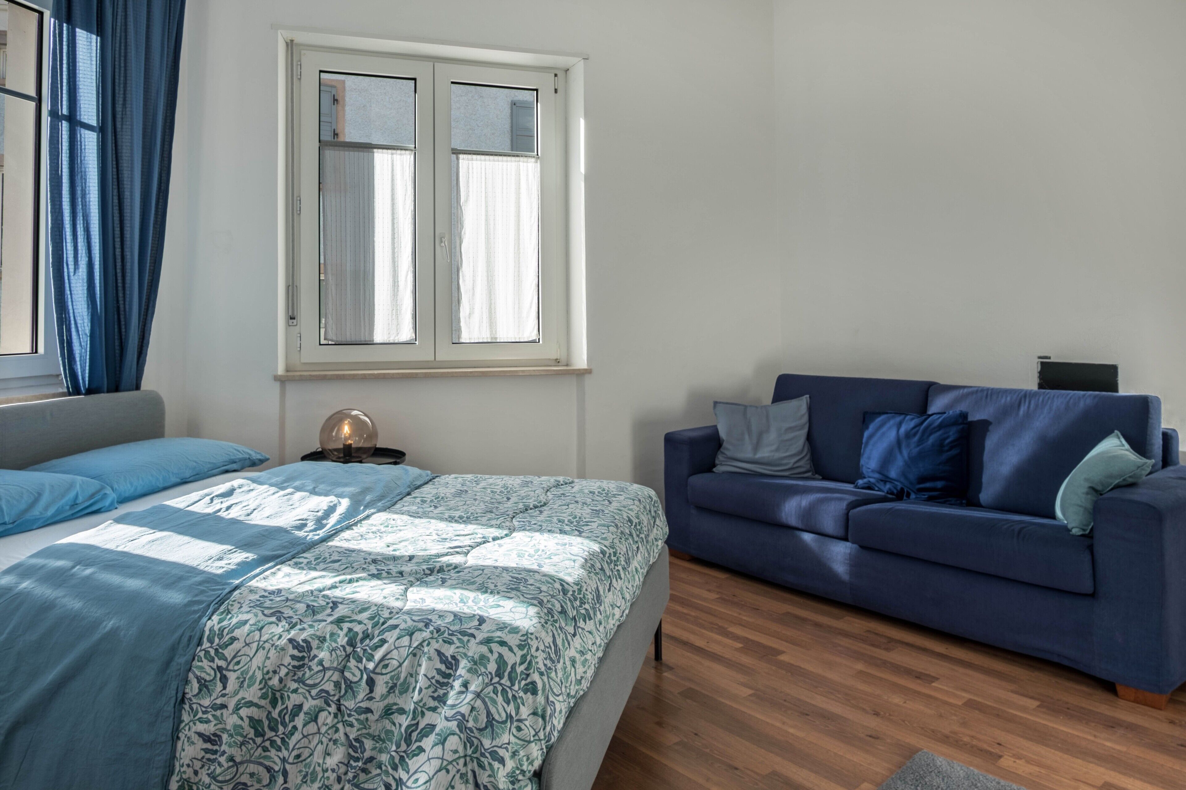 Quarto com cama, sofá azul, janela com cortinas e mesa de cabeceira com abajur.