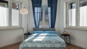 Un dormitorio con una cama, dos mesitas de noche, una lámpara de pie y un ventanal con cortinas.