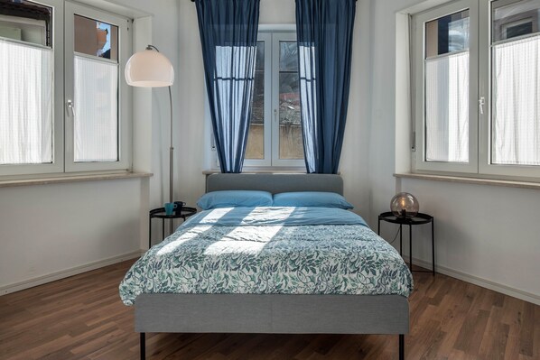 A bedroom with a bed, two bedside tables, a floor lamp, and a window with curtains.