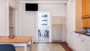 Una cocina pequeña con armarios blancos, una mesa de madera y un televisor sobre la puerta.