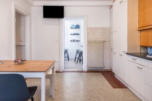 Una cuina compacta amb armaris blancs, una taula de fusta i un televisor fixat sobre la porta.