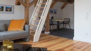 A loft-style room with exposed wooden beams, a white ladder, a gray sofa with an orange cushion, a small table with two chairs, and a projector on a desk. - Apartment 1 Wohnen am Turm - Living at Schloßplatz (Oldenburg)