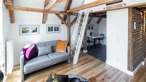 A modern living room with a grey sofa, wooden beams, a white ladder, and a dining area with a table and chairs. - Apartment 1 Wohnen am Turm - Living at Schloßplatz (Oldenburg)