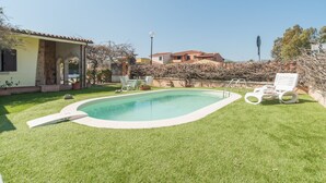 Una piscina con una pequeña terraza, rodeada de un césped cuidado y una casa con tejado de baldosas.