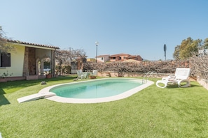 En swimmingpool med en lille terrasse, omgivet af en velholdt græsplæne og et hus med tegltag.