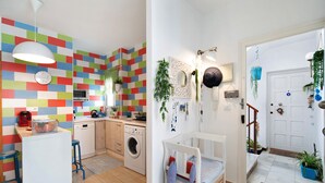 A kitchen with a washing machine, a dining area with a table and chairs, and a small white bed in a hallway. - Apartment Caepionis close to the Beach with Shared Rooftop Terrace & Wi-Fi (Chipiona)