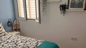 A bedroom with a bed, a window with blinds, a surfboard, a plant, and a framed picture on the wall. - Apartment Caepionis close to the Beach with Shared Rooftop Terrace & Wi-Fi (Chipiona)