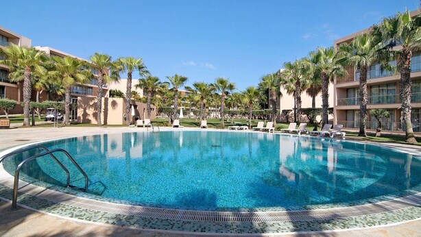 A large, clear swimming pool surrounded by palm trees and lounge chairs, with modern buildings in the background. - Apartamento Salgados Sol e Sal with Pool, Wi-Fi, A/C & Garden (Guia)