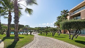 A paved pathway leading to a pool area with palm trees and a building in the background. - Apartamento Salgados Sol e Sal with Pool, Wi-Fi, A/C & Garden (Guia)