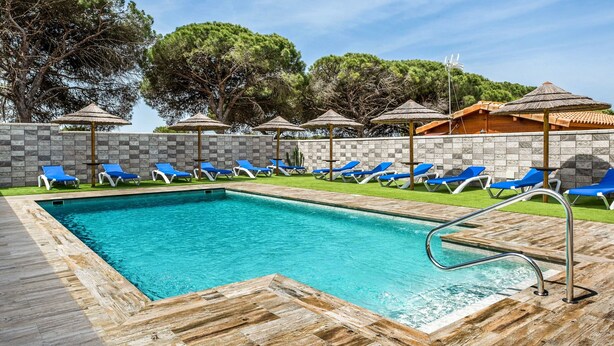 Piscina con sillas de descanso azules y sombrillas, rodeada por un muro de piedra y árboles.