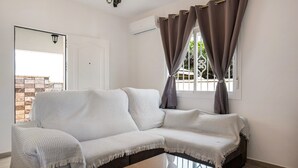 Una habitación con un sofá blanco, una mesa de centro de vidrio y una ventana con cortinas.