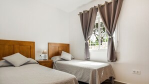 Una habitación con dos camas, cabeceros de madera, una mesita de noche con lámpara y una ventana con cortinas.