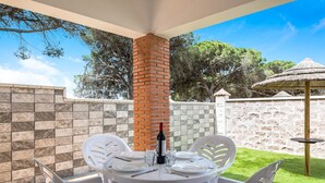 Un patio con una mesa redonda, sillas de plástico blanco, una botella de vino y una pequeña mesa con una bebida.