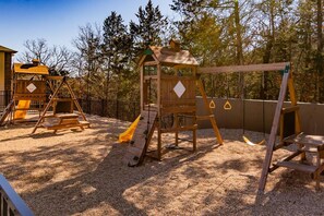 Une aire de jeux avec des structures en bois et un toboggan, entourée d’arbres.