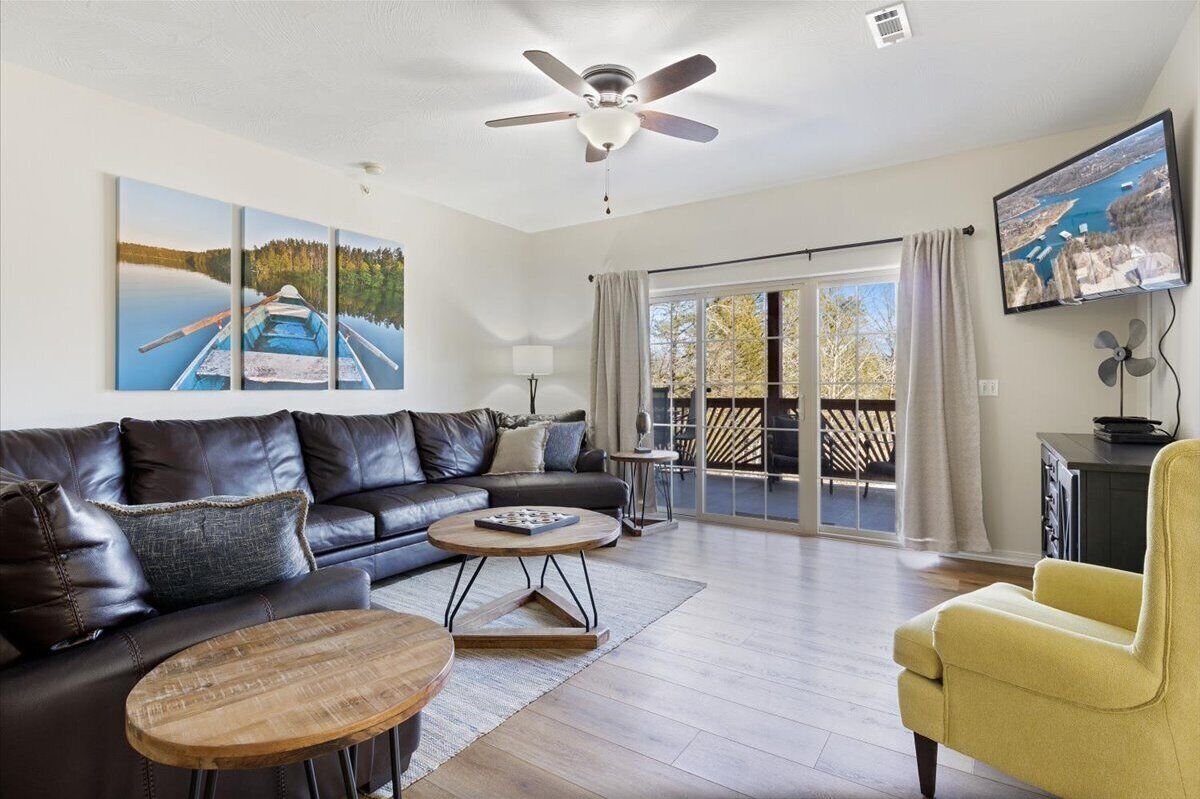 A modern living room with a leather sofa, a yellow armchair, and a wooden coffee table. There is a ceiling fan, a flat-screen TV mounted on the wall, and a large window with curtains.