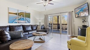 Un salon moderne avec un canapé en cuir, un fauteuil jaune et une table basse en bois. On y trouve un ventilateur de plafond, un téléviseur à écran plat fixé au mur et une grande fenêtre avec des rideaux.
