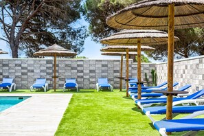 Zona de piscina con sillones reclinables azules, sombrillas de paja y un muro de piedra.