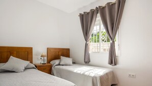 A bedroom with two beds, a nightstand with a lamp, a window with curtains, and a view of greenery outside. - Cozy Casa Torre de Meca 1 with Pool, Terrace, and Garden (Los Caños de Meca)