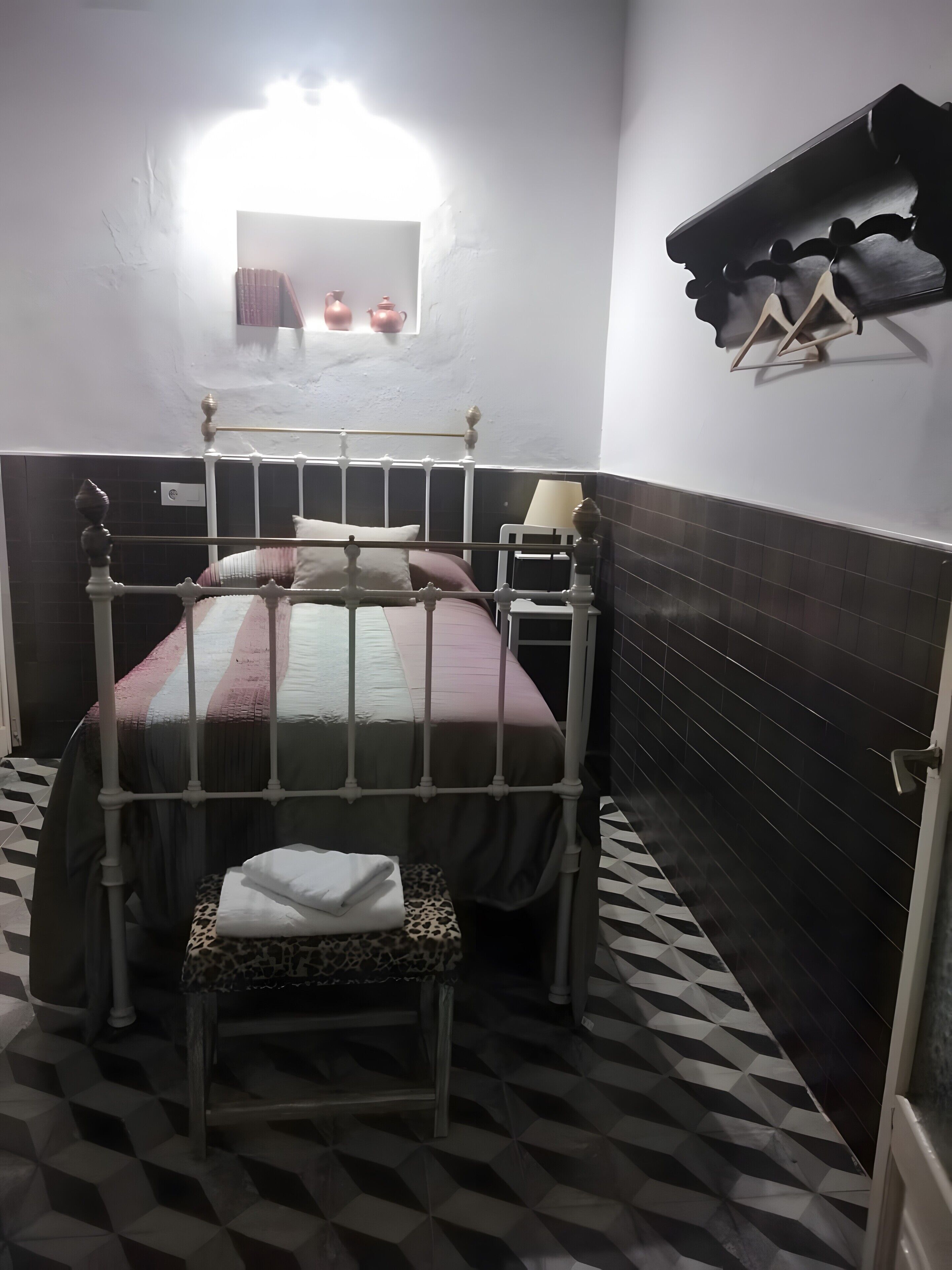 Una habitación pequeña y tenuemente iluminada con una cama individual, una mesa pequeña y una silla. El piso tiene un estampado de cuadros y una pared oscura con un estante sobre la cama.