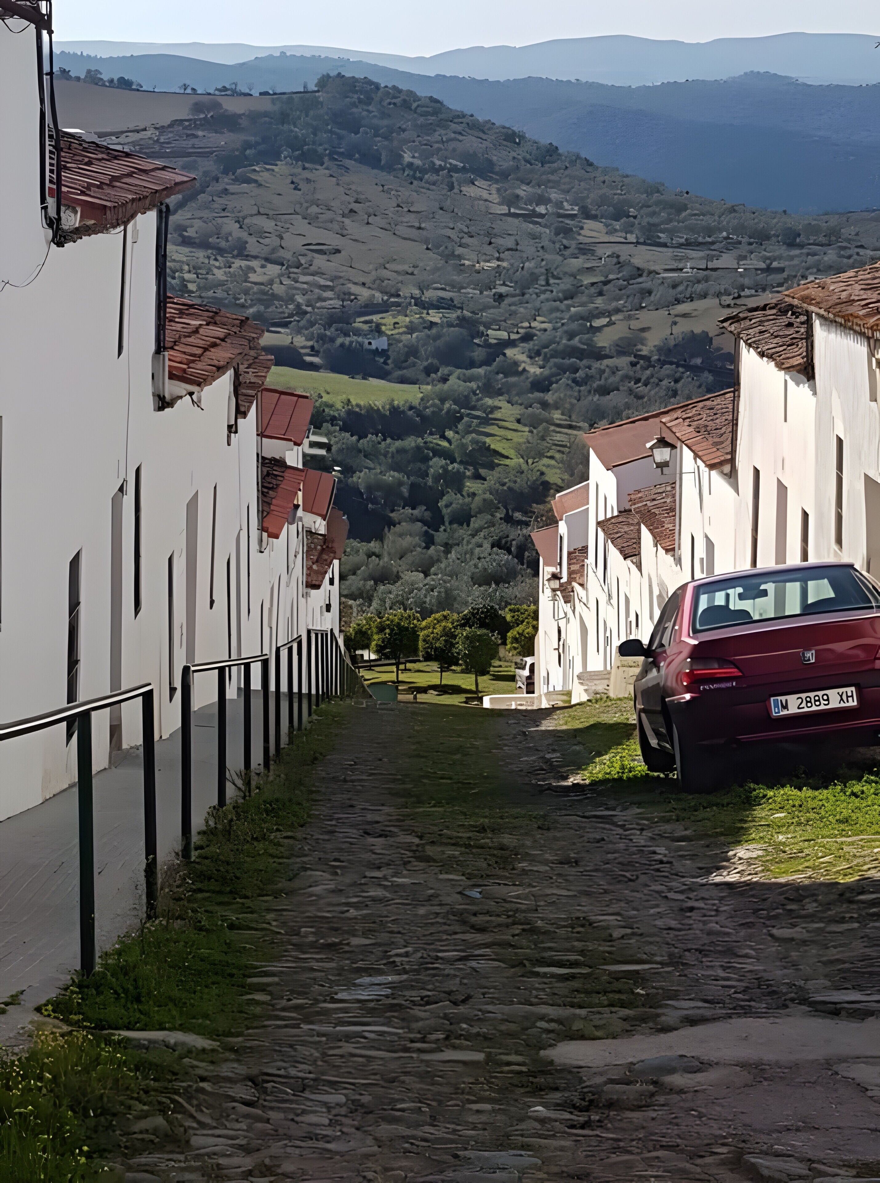 A narrow cobblestone street flanked by white buildings with red-tiled roofs, leading to a hilly landscape.