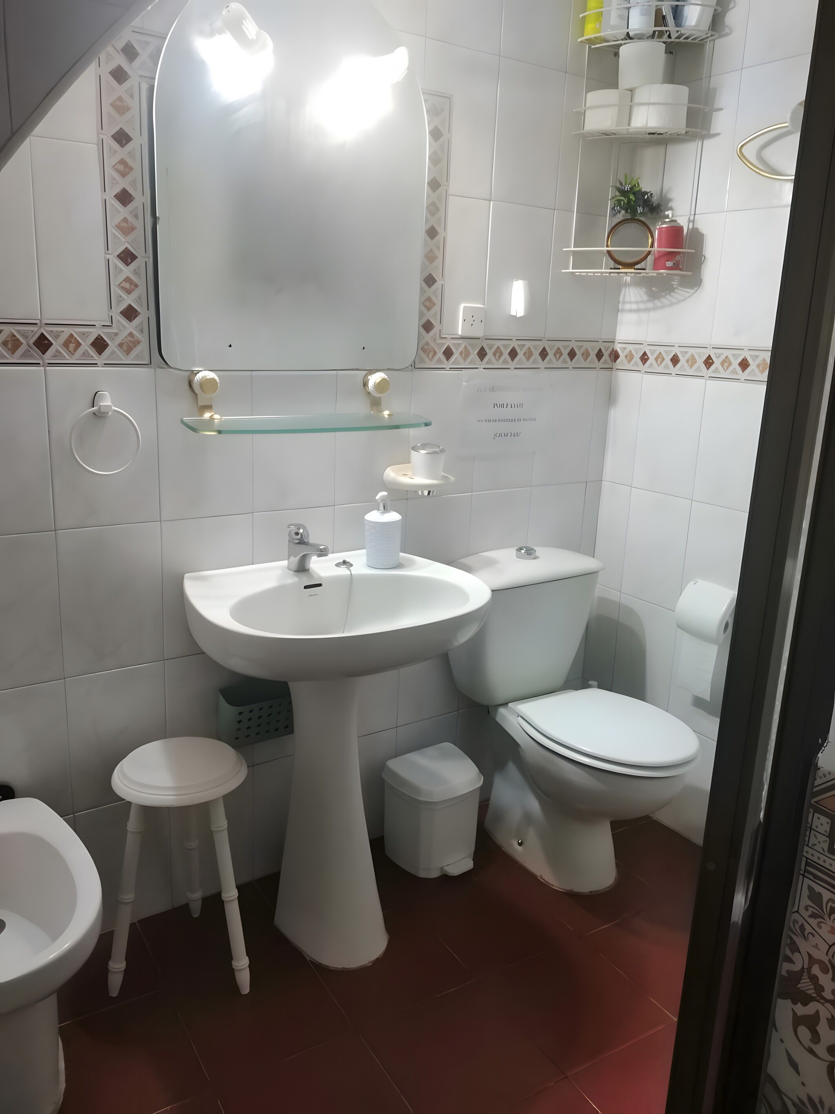 Un baño con lavamanos, inodoro y espejo. Hay estantes con artículos de aseo personal y un taburete pequeño.