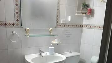 Un baño con lavamanos, inodoro y espejo. Hay estantes con artículos de aseo personal y un taburete pequeño.