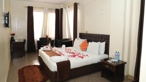 A hotel room with a bed, a desk, a chair, a small table, and a brown rug. - Goshen Inn Eldoret (Eldoret)