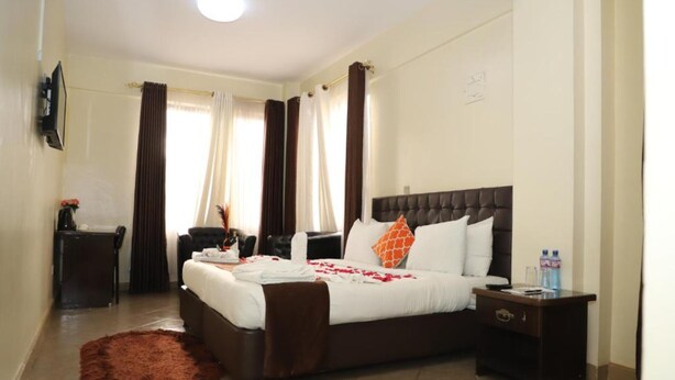 A hotel room with a large bed, a nightstand, a TV mounted on the wall, and a window with curtains. - Goshen Inn Eldoret (Eldoret)