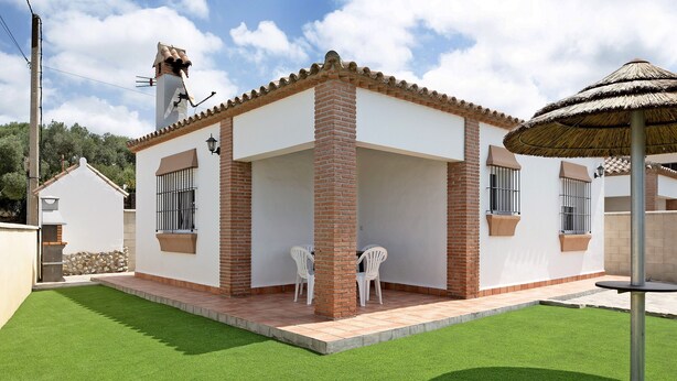 A house with a patio, two white chairs, and a thatched umbrella. - Charming Casita Torre de Meca 1 with Terrace, Garden, and Wi-Fi (Los Caños de Meca)