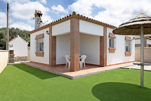 Une maison avec un patio, deux chaises blanches et un parasol en chaume.