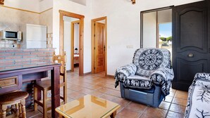 A living room with a brick fireplace, wooden furniture, a patterned armchair, and a dark door. - Charming Casita Torre de Meca 1 with Terrace, Garden, and Wi-Fi (Los Caños de Meca)