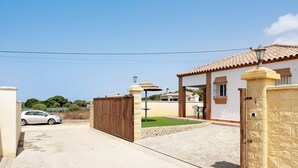 A gated residential area with a paved driveway, a parked car, and a house with a tiled roof. - Charming Casita Torre de Meca 1 with Terrace, Garden, and Wi-Fi (Los Caños de Meca)
